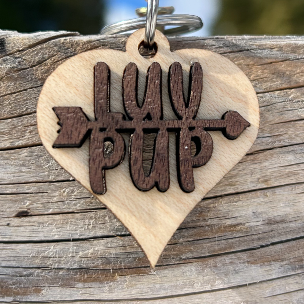 Solid maple wood heart collar charm, with solid walnut wood inlay with text that says LUV on top of the text PUP and an arrow going between the two words. it is attached to a spit ring